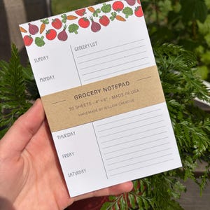 May include: A white paper grocery notepad measuring 10.16 cm by 15.24 cm with a brown paper belly band. The top features a colourful vegetable pattern of tomatoes, carrots, broccoli, and onions. The layout includes sections for days of the week.