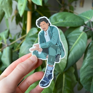 Könnte beinhalten: Ein Aufkleber mit einer Illustration einer Person in einem tealfarbenen Anzug und Stiefeln. Die Person sitzt mit einem Bein über dem anderen. Der Hintergrund ist verschwommenes grünes Laub.