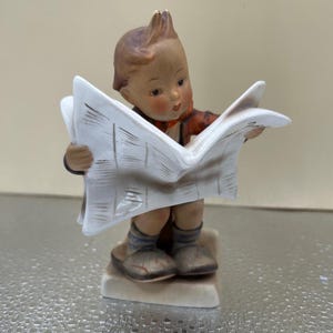 May include: A porcelain figurine of a boy reading a newspaper. The boy is wearing a red and white shirt and blue pants. He is sitting on a small base.