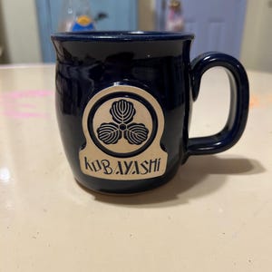 Może przedstawiać: Kubek ceramiczny w ciemnoniebieskim kolorze z złotym emblematem przedstawiającym stylizowany tr&oacute;jlistny koniczyna i napis "KUBAYASHI".
