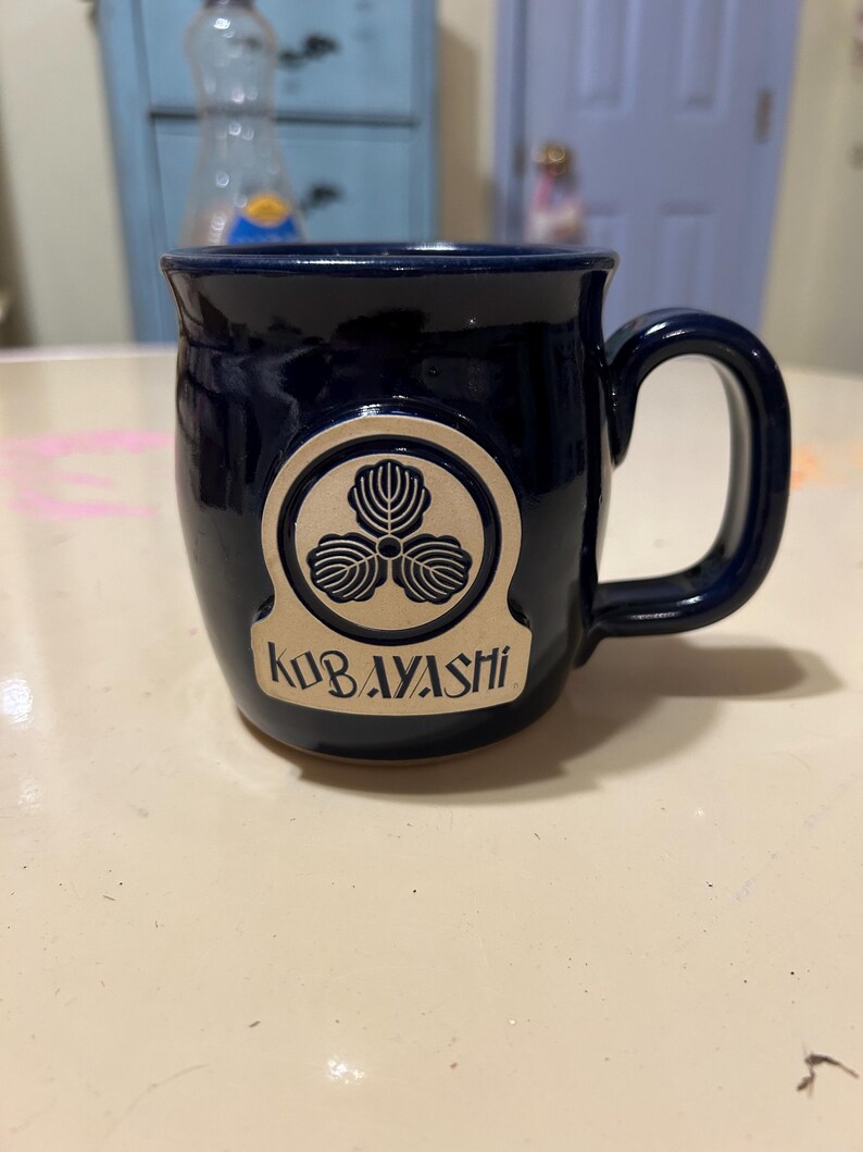 Może przedstawiać: Kubek ceramiczny w ciemnoniebieskim kolorze z złotym emblematem przedstawiającym stylizowany tr&oacute;jlistny koniczyna i napis "KUBAYASHI".