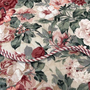 May include: A quilted fabric featuring a floral pattern with large red, pink, and white roses set against a beige background. The material includes green leaves and a decorative red and white striped piping trim along the folded edge.