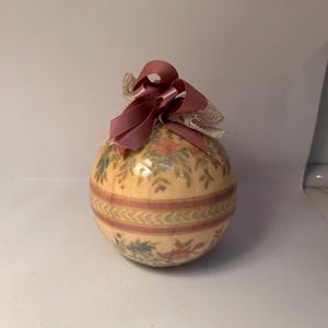 May include: A spherical ornament featuring a cream-colored base with a floral pattern in shades of pink, green, and blue. A horizontal striped band wraps around the center. The top is adorned with a dusty rose ribbon bow and white lace trim.