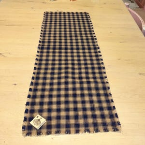 May include: A rectangular table runner with a navy blue and tan plaid pattern. The edges are fringed. A small tag with text is attached to the corner of the runner. The runner is displayed on a light-colored wooden table.