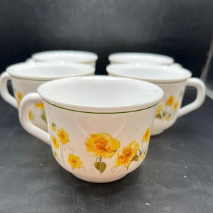 May include: Set of white ceramic teacups with a floral design featuring yellow flowers and green accents. Each cup has a handle and a green trim around the rim. The cups are arranged on a dark surface.
