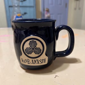 Może przedstawiać: Kubek ceramiczny w ciemnoniebieskim kolorze z złotym logo przedstawiającym stylizowany tr&oacute;jlistny koniczyna i napis "Kobayashi".