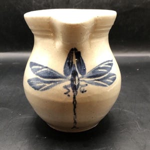 May include: A small, cream-colored ceramic pitcher with a blue dragonfly design. The pitcher has a rounded body and a flared lip.