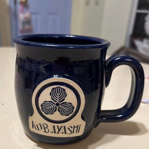 Może przedstawiać: Kubek ceramiczny w ciemnoniebieskim kolorze z złotym logo przedstawiającym koniczynę tr&oacute;jlistną i tekstem "Kobayashi".