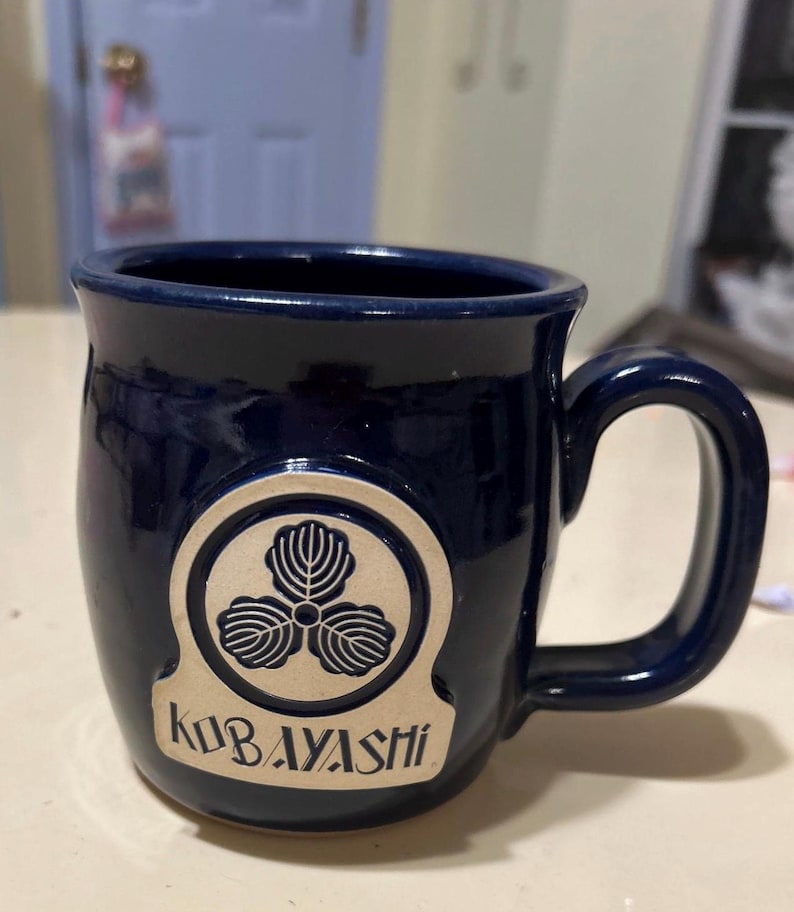 Może przedstawiać: Kubek ceramiczny w ciemnoniebieskim kolorze z złotym logo przedstawiającym koniczynę tr&oacute;jlistną i tekstem "Kobayashi".