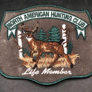 May include: A brown and green embroidered patch with a white banner that reads "North American Hunting Club". The patch features a white-tailed deer standing in a forest setting with the words "Life Member" below the deer.