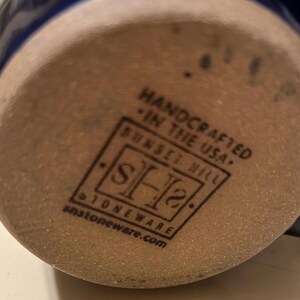 Może przedstawiać: Zbliżenie na sp&oacute;d kubka z kamionki. Kubek jest brązowy z niebieską glazurą. Na spodzie kubka nadrukowano słowa "Handcrafted in the USA." i "Sunset Hill Stoneware.com".