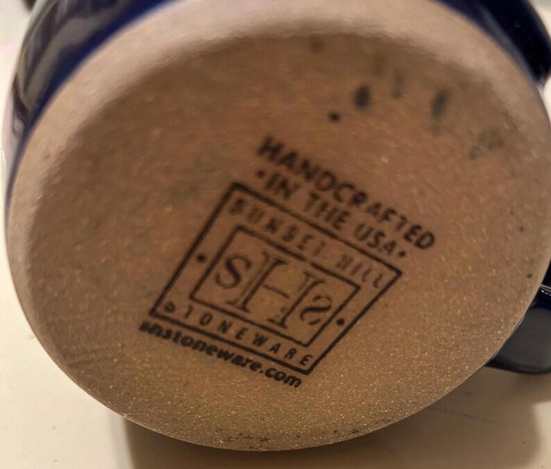 Może przedstawiać: Zbliżenie na sp&oacute;d kubka z kamionki. Kubek jest brązowy z niebieską glazurą. Na spodzie kubka nadrukowano słowa "Handcrafted in the USA." i "Sunset Hill Stoneware.com".
