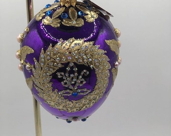 May include: Purple glass Christmas ornament with gold and blue embellishments. The ornament is decorated with a gold wreath and a blue and gold floral design. The ornament is hanging from a gold hook.