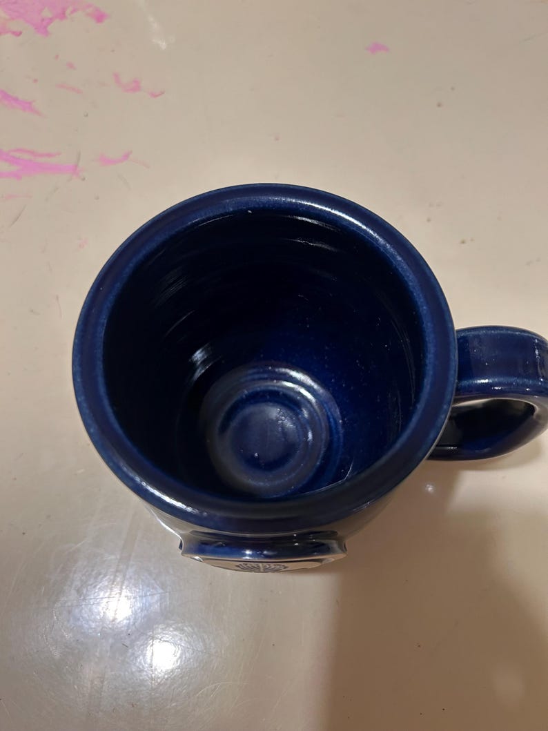 Może przedstawiać: Kubek ceramiczny w ciemnoniebieskim kolorze z srebrną obw&oacute;dką i srebrnym logo na spodzie.