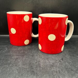 May include: Two red ceramic mugs with white polka dots. Each mug has a white handle and a white interior. The mugs are cylindrical in shape and sit on a dark surface.