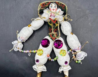 May include: A white porcelain clown doll with a green, red, and gold patterned costume. The doll has a white lace collar and cuffs, and is decorated with pink and yellow rhinestones. The doll is hanging from a gold string.
