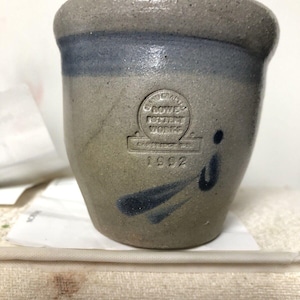 May include: A small, gray stoneware crock with a blue glaze. The crock has a stamped logo that reads "Bowles Pottery Works, Cambridge, MA" and the year "1992".