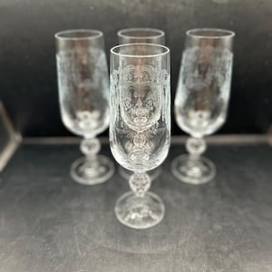 May include: Four clear champagne flutes with ornate etched designs. Each glass features a detailed face-like pattern near the bowl and a decorative stem. The glasses are arranged on a reflective surface, showcasing their elegant design.