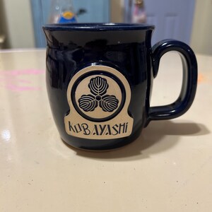 Może przedstawiać: Kubek ceramiczny w ciemnoniebieskim kolorze z złotym emblematem przedstawiającym stylizowany tr&oacute;jlistny koniczyna i napis "KOBAYASHI".