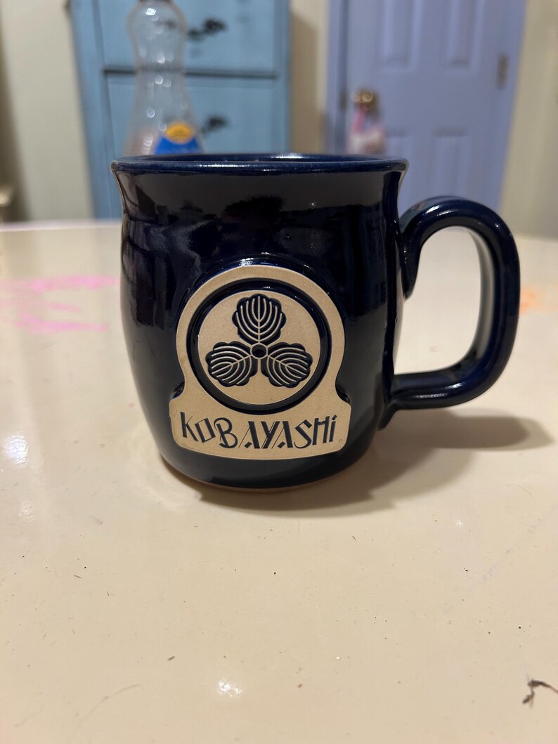 Może przedstawiać: Kubek ceramiczny w ciemnoniebieskim kolorze z złotym emblematem przedstawiającym stylizowany tr&oacute;jlistny koniczyna i napis "KOBAYASHI".