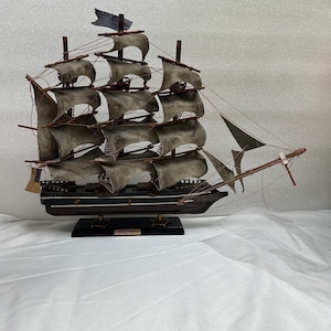 May include: A detailed wooden model of a tall ship with multiple sails and rigging. The ship has a dark hull, tan sails, and a small blue flag at the top of the mast. The model sits on a black base.