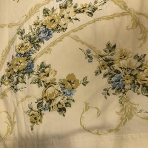 May include: Cream-colored fabric with a floral pattern featuring yellow, blue, and green flowers. The design includes decorative, light green, curved lines. The fabric has a soft, slightly wrinkled appearance.