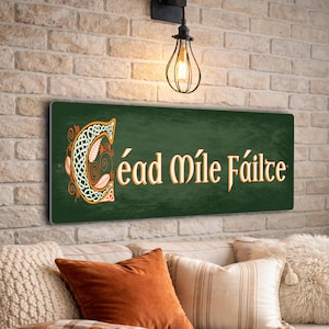 Può includere: Un cartello rettangolare verde con la scritta "Céad Míle Fáilte" in lettere dorate. La lettera "C" è elaboratamente progettata con nodi celtici arancioni e bianchi. Il cartello è montato su un muro di mattoni sopra un divano con cuscini.