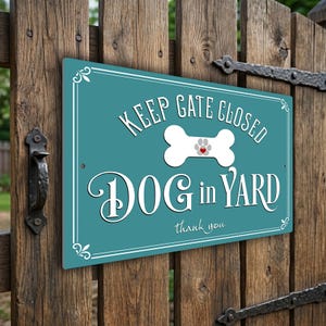 May include: A teal metal sign with white text saying "KEEP GATE CLOSED DOG in YARD thank you." A white bone with a paw print and heart is in the center. The sign is mounted on a wooden gate with metal hinges.