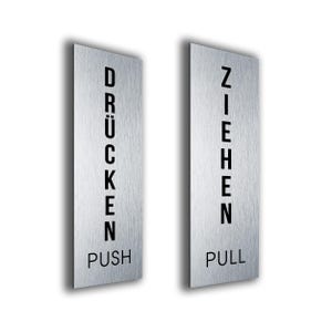 Könnte beinhalten: Zwei Türschilder aus gebürstetem Metall. Auf einem Schild steht "DRÜCKEN" über "PUSH" in schwarzer Schrift. Auf dem anderen Schild steht "ZIEHEN" über "PULL" in schwarzer Schrift. Die Schilder sind rechteckig und leicht schattiert.