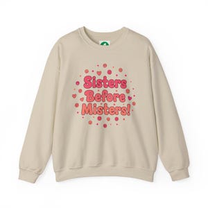 Puede incluir: Sudadera beige con el texto "Sisters Before Misters!" en rosa y rojo. El texto está rodeado de corazones y caras sonrientes. La sudadera tiene mangas largas y cuello redondo.