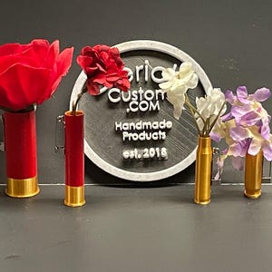 May include: Three bullet shell vases with flowers. The vases are painted red and gold. The flowers are red, white, and purple. The sign behind the vases reads "Brick Custom .COM Handmade Products est. 2018".