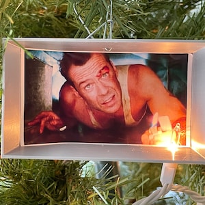 May include: A rectangular Christmas tree ornament featuring a movie still of a man with a bloody forehead. The image is set in a silver frame and hangs from a silver wire. A small light shines from the bottom.
