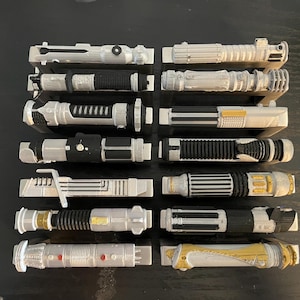 May include: Collection of toy lightsabers in various designs, primarily silver and black, with some gold accents. The lightsabers are arranged in two columns, showcasing their detailed handles and emitter designs. The overall theme is science fiction.
