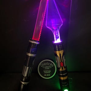 May include: Two custom cake servers with glowing blades. One is a serrated knife, the other a triangular server. The handles are black and gold, with silver accents. The blades glow red and purple, with green and white lights at the base.