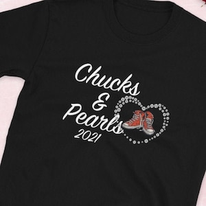 May include: Black t-shirt with white text that reads "Chucks & Pearls 2021". A pair of red sneakers with white laces are inside a heart made of white rhinestones.