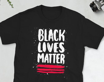 Puede incluir: Camiseta negra con las palabras "Black Lives Matter" en letras blancas con rayas rojas debajo. La camiseta está sobre una superficie blanca.