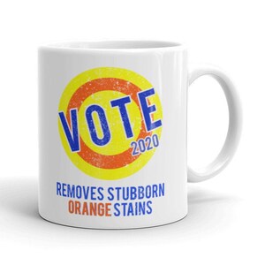 May include: White ceramic mug with a yellow and orange circle design that says "VOTE 2020" in blue letters. The text below the circle says "REMOVES STUBBORN ORANGE STAINS".