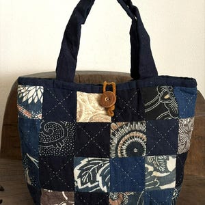 Könnte beinhalten: Eine Patchwork-Tragetasche mit marineblauem Äußeren und Griffen. Die Tasche hat eine Vielzahl von gemusterten Quadraten in Blau-, Creme- und Brauntönen. Ein Holzknopf sichert die Oberseite. Die Tasche steht auf einer Holzoberfläche.
