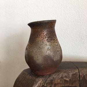 以下が含まれることがあります： 素朴で質感のある仕上がりの手作りのセラミック花瓶。花瓶は、濃い茶色のトップから、まだらの薄茶色と灰色のミドルセクションにフェードし、赤茶色のベースに移行します。 花瓶はわずかにフレアした開口部を持っています。