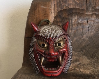 May include: A red and silver Japanese demon mask with a wide open mouth and sharp teeth. The mask is made of wood and has a traditional design.