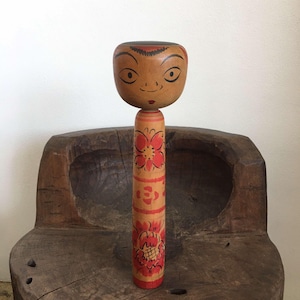 Puede incluir: Una muñeca de madera japonesa tradicional con la cara pintada y diseños florales. La muñeca está de pie sobre una superficie de madera.