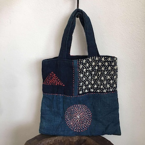 May include: A patchwork tote bag with a dark blue denim base. The bag features a red triangle and a spiral design, both stitched with red thread. The top section has a white and black dotted pattern.