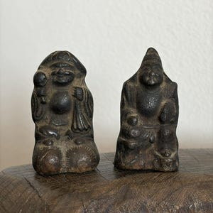 May include: Two small, dark-colored statues of seated figures. The statue on the left has a smiling face and holds a round object. The statue on the right has a pointed hat and is seated with its hands on its knees. Both statues are on a wooden surface.