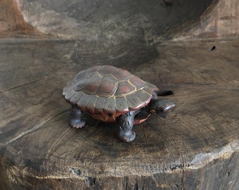 May include: A small, intricately carved wooden turtle figurine with a reddish-brown shell and black legs. The turtle is sitting on a rough, wooden surface.