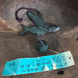 Könnte beinhalten: Ein grüner Windspiel aus Metall mit einem fliegenden Fisch-Design, einer Glocke und einem blauen und weißen Etikett mit japanischem Text, der "Hoch fliegen, weit fliegen, frei fliegen" lautet.