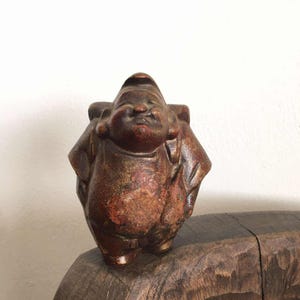 May include: A small, brown ceramic statue of a smiling, rotund figure with a traditional hat. The statue has a reddish-brown hue with gold accents and is likely a decorative item or collectible.
