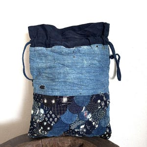 Può includere: Un sacchetto con coulisse in patchwork di denim. La parte superiore è blu scuro, il corpo è in denim blu chiaro e scuro, con cuciture visibili. La sezione inferiore presenta tessuti fantasia nei toni del blu.