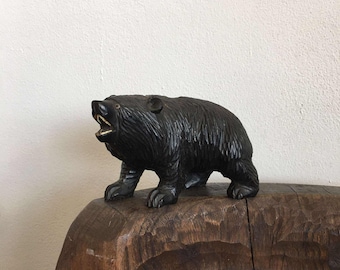 May include: A carved black bear figurine with an open mouth, revealing a gold-colored interior. The bear is standing on all fours, with detailed fur texture. The figurine is placed on a carved wooden surface.