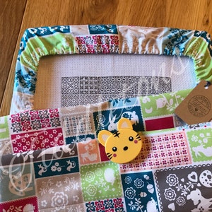 May include: A white embroidery hoop with a colourful fabric cover. The fabric has a patchwork design with various animals and floral patterns. A yellow tiger-shaped needle minder is on the fabric. The hoop is on a brown wooden surface.