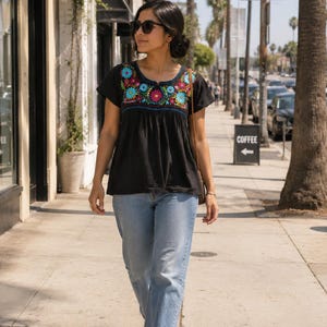May include: A black short-sleeved blouse featuring a vibrant floral embroidery pattern in shades of blue, red, and yellow around the neckline, paired with light blue denim jeans and white trainers. The blouse has a loose, gathered fit.